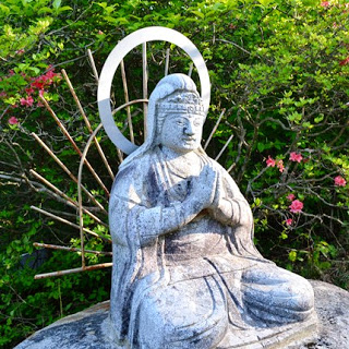 ツツジの緑に囲まれ、岩の上に正座し、円形の後光を背負った観音像が合掌している写真
