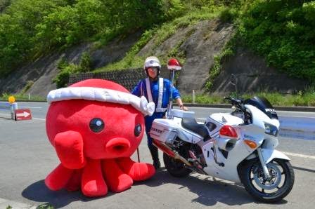 白バイの警察官がオクトパス君の着ぐるみと並んで立ち、背景に山道が見える場所で撮影されている記念写真