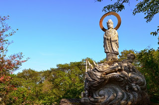雲一つない青空を背景に、ツツジや木々に囲まれたなだらかな斜面に、 龍のような彫刻の上に立つ、円形の光背を背負った観音像の写真