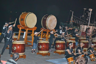 夜の港の岸壁で、大小さまざまな和太鼓を並べて、子どもから大人までが法被姿で力強く太鼓を打ち鳴らしている和太鼓演奏の写真