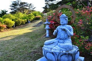 木々やツツジに囲まれ、石灯篭が設置された緩やかな斜面沿いに座っている、 線香を持つ観音像の写真