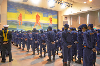 会場内で紺色の制服を着た多くの消防団員が手を後ろで組み、整列している後ろ姿を写した写真