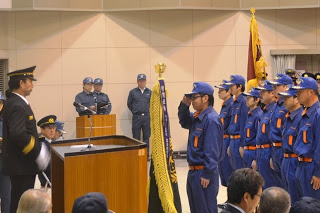 制服と制帽を着た男性と紺色の制服を着た消防団員が演台を挟んで向かい合い、敬礼している式典の様子の写真