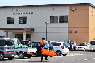 駐車場に沢山の車が停まっている公立南三陸診療所の前で、青い制服の救急隊員がオレンジ色の担架を持って歩いている写真