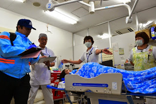 医師と看護師たちがベッドに横たわる患者の容態を救急隊員に伝えている訓練の様子の写真