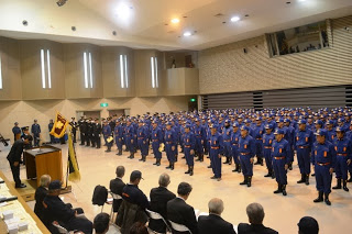 紺色の制服を着た多数の消防団員が整列して並び、演題の前で会釈している男性や来賓の方々が前方の椅子に並んで座っているなど式典会場の全体の様子を写した写真