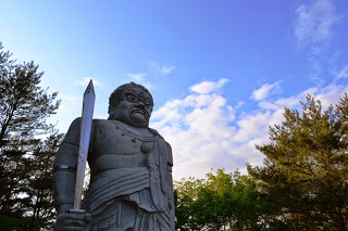 背景には木々と雲が少し浮かぶ空が広がり、剣を持った大聖不動明王をアップで撮影した写真
