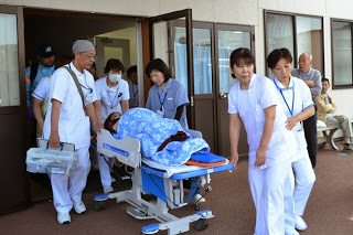 青い毛布をかけられた患者をのせたベッドを医師と看護師たちが連携して屋外に搬送している写真