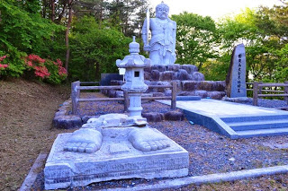 石灯籠や文字が刻まれた石碑と、階段付きの台座の上に立つ剣を持った大聖不動明王が柵の中に設置され、手前には震災で倒壊した足の石造物がある写真