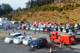 複数の消防車と関係者の車が駐車場に並び、周囲に消防団員や関係者がいる駐車場の様子を写した写真