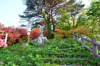 手すりのついた階段を上がった先に、赤やピンク色の椿の花や木々に囲まれた斜面に設置された灑水観音様の写真
