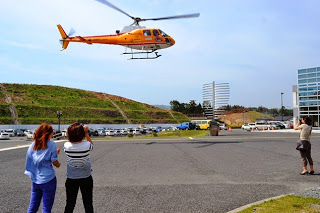 病院前で二人の女性がカメラやスマートフォンを構えて、離陸するヘリコプターを見送っている写真