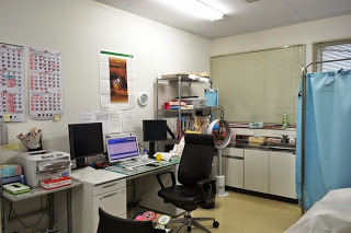 デスクにパソコンやプリンターが置かれていて、周囲には薬品や医療器具が収納されている棚があり、窓のある清潔な部屋に診察用のベッドとカーテンも見える診察室の写真