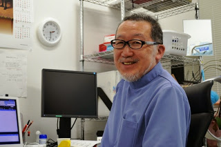 眼鏡をかけてひげを生やした男性医師が、パソコンのあるデスクに座って笑顔でカメラを向いており、後ろの棚に医療器具や日用品が置かれている診療所内の医師の写真