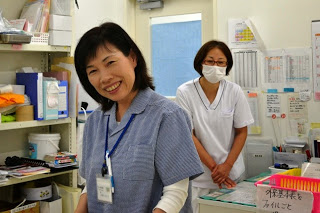 手前に青いユニフォームを着た女性スタッフが笑顔で立ち、奥には白衣を着てマスクをした別の女性スタッフが立っており、棚や掲示物のある診療所の受付または事務スペースの写真