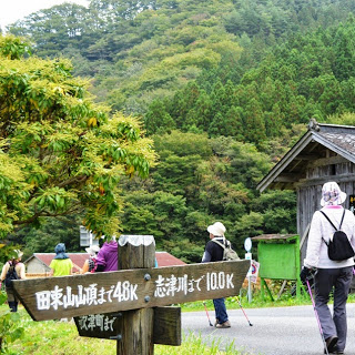 山道の分岐点にある木の看板に「田束山山頂まで4.8キロ」「志津川まで10.0キロ」と書かれており、数人の参加者が周囲を歩いている様子が写っている写真