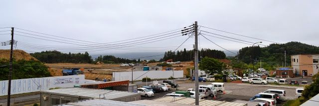 重機が動いている大規模な造成地と、手前にある駐車場やプレハブの建物が並ぶ写真