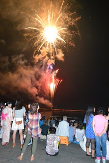浴衣や私服姿の人々が海辺の防波堤に座ったり立ったりして夜空を見上げ、目の前で上がる色とりどりの花火を楽しんでいる様子を後方から撮影した写真