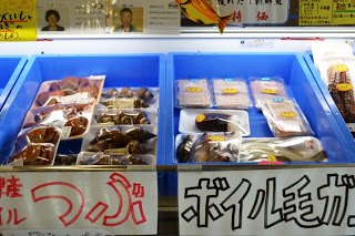 左にはつぶ貝、右にはボイル毛ガニと書かれた手書きの表示がされた青い保冷ケースに真空パックされた海産物が整然と並べられている写真