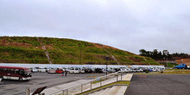 盛り土された斜面に草が生え、手前の広い駐車場にはたくさんの車が停まっている写真