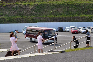 斜面のある駐車場に赤い送迎バスが停まり、階段の途中に立ち止まっているピンクの制服を着た看護師たちがヘリコプターを見送っている写真