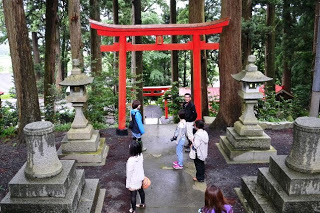 森の中にある神社の赤い鳥居の前で、数人の参加者が石灯籠に囲まれて立ち止まっている写真