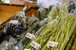 店内のかごの中に、袋詰めされたナス、キュウリ、空芯菜などの新鮮な野菜が価格シール付きで並べられている写真