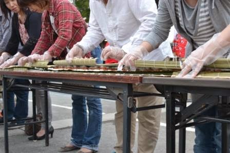 人々が両手にビニール製の手袋をつけて、長い巻き寿司を一斉に巻いている様子の写真