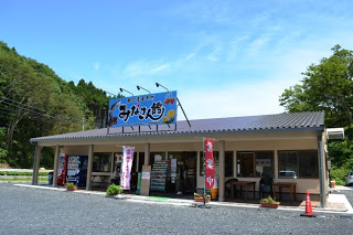 木々に囲まれた郊外の広場に建つ平屋の建物の正面に「みなさん館」大きく書かれた青い看板が掲げられ、軒下には幟旗や机、いすが並ぶ南三陸直売所 みなさん館の外観写真