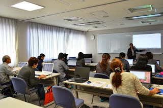 窓際にカーテンが引かれた明るい教室で、大人たちがパソコンを開いて机に向かい、前方の講師がホワイトボードの前に立って話をしている様子の写真