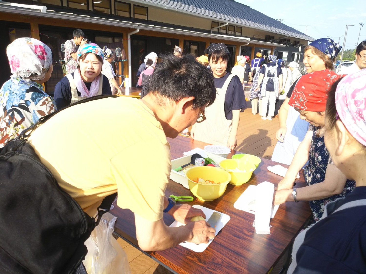 屋外のテーブルで複数の人たちがエプロンと三角巾をつけて食材を切ったり話したりして、調理の準備をしている写真