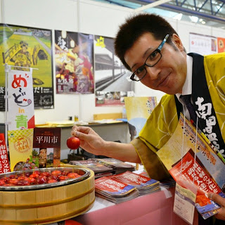 黄色い法被を着た男性が、赤い実が入った桶の前でその一粒を持ち上げながら笑顔で紹介している観光PRブースの写真