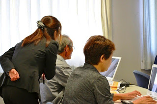 女性講師が高齢の男性にパソコンの操作を丁寧に教えていて、その横で別の女性も画面を見ながら作業している写真
