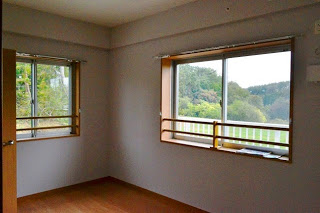 白壁でフローリング床の角部屋にある2つの窓から、外の森や山が見える様子が写っている写真