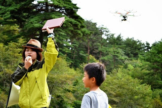 広場で男の子が横で見ているなか、帽子を被った女性が拡声器で説明しながらファイルを持った手を掲げた右上の上空にドローンが飛んでいる写真