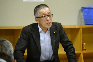 和室でスーツを着た眼鏡をかけた男性が、座った状態で真剣な表情で発言している様子が写っている写真