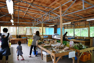 木造の直売所内で、親子連れを含む客が、木の台にならんだ野菜や加工品の品物を見ている店内の様子を写した写真