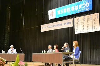 「南三陸町福祉まつり」の横断幕が掲げられたステージで、進行役の男性と、4人の登壇者が座ってパネルディスカッションをしている写真