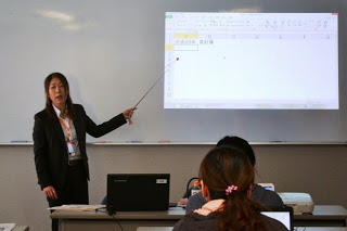 黒いスーツ姿の女性講師が指示棒を使ってホワイトボードのスクリーンに映されたExcelの画面を説明している写真