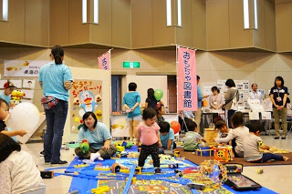 「おもちゃ図書館」と書かれたのぼり旗のスペースで、子どもたちが床に敷かれたプラレールを見たり、おもちゃで楽しそうに遊んでいる写真