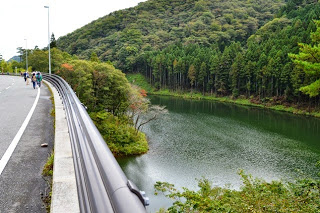 ガードレール越しに緑に囲まれた静かな川と杉林が広がる舗装された橋を、数人の参加者が歩いている写真