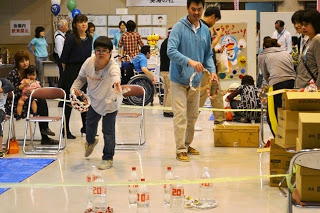 大人たちが見守っているなか、屋内でペットボトルを的にした輪投げに男の子が挑戦している写真