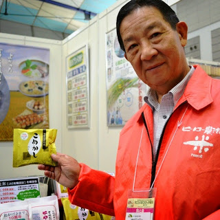 赤いジャンパーを着た男性が、「こめかし」と書かれた袋入りのお菓子を笑顔で持ちながら紹介している様子を写した物産展の写真