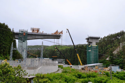 深い森の谷間に巨大な橋の支柱が立ち、クレーンで橋が建設されている途中の写真