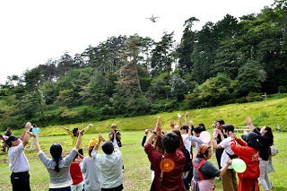木々に囲まれた広場で、大勢の人が空に向かって手を挙げ、飛んでいるドローンに向かって声援を送っている写真