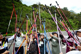 帽子をかぶった参加者たちが集まり、色とりどりのトレッキングポールを元気よく空に向かって高く突き上げている様子が写っている写真