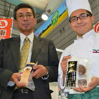 スーツ姿の男性とコック帽をかぶったパティシエが、それぞれ地元の特産菓子を手に持ち、カメラに向けてアピールしている写真