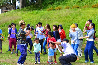草原のような広場で、子どもたちと大人たちが並んで笑いながら踊ったりポーズを取ったりしている様子を、カメラを持った男性が撮影している写真