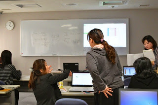 グレー色の上着を着た女性が自席から立ち上がり、パソコンの右側に跪いて座っている講師に質問を行っている写真