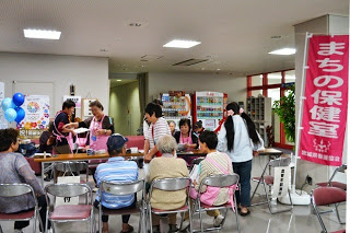 「まちの保健室」と書かれたピンクののぼり旗の傍で、エプロン姿のスタッフが椅子に座る高齢者の健康相談を行っている写真